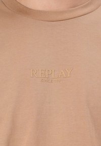 Bež majica iz bombaža s rebrastim vratnim izrezom in vtisnjenim logotipom "REPLAY SINCE 1981" v usklajeni barvi. Mehka, gladka tekstura.