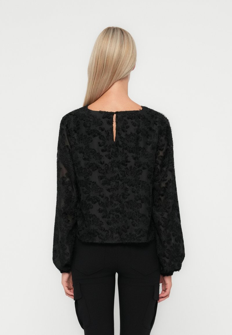 Czarna bluzka z teksturowanym kwiatowym wzorem, przezroczystym materiałem i puffowanymi rękawami. Detal w kształcie dziurki z tyłu, krótki fason. Połączona z czarnymi spodniami.