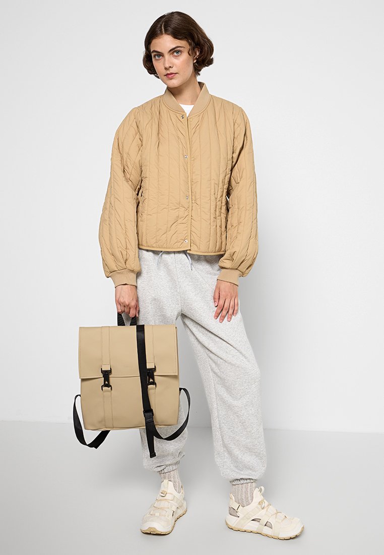 Giacca bomber trapuntata beige, pantaloni da jogging grigi chiari, zaino beige con spallacci regolabili e sneakers bianche. Design semplice e moderno.