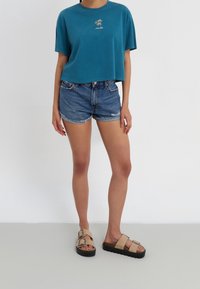 Teal cropped T-shirt met geborduurde palmboom, gecombineerd met denim shorts en beige open-toe sandalen met dubbele gespen en zwarte zolen.