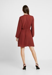 Vestido burdeos con mangas largas, cintura ajustada y borde de encaje en el escote. Tela fluida y longitud por encima de la rodilla, combinado con tacones negros.