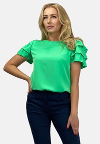 Zielona bluzka z krótkimi, warstwowymi rękawami w falbanki, gładka tekstura, okrągły dekolt, połączona z ciemnoniebieskimi spodniami, dopasowany fason.