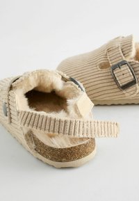 Beige slip-on skor med ribbat tyg, fluffig innersula, justerbart band och korksula. Texturerad detaljering och silverspänne som accent.