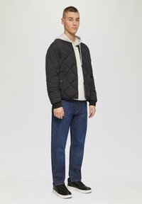 Veste bomber matelassée noire avec capuche sur un sweat à capuche gris clair, associée à un jean bleu et des baskets noires, affichant un look décontracté.