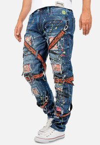 Jean en denim usé avec un tissu bleu et des éclaboussures de peinture colorées. Détails notables incluant plusieurs pièces, sangles et poches.
