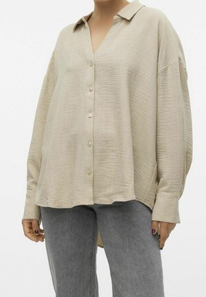 Luźna beżowa teksturowana koszula na guziki z długimi rękawami, zestawiona z szarymi dżinsami, pokazana na osobie przyciętej na wysokości szyi i kolan.