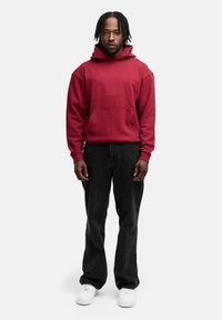 Rote Kapuzen Sweatshirt mit einer Tasche auf der Vorderseite, kombiniert mit schwarzen Jeans und weißen Sneakers. Das Model steht aufrecht mit entspannten Armen.