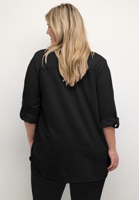 Maglietta nera a maniche lunghe con polsini arrotolati, texture morbida e orlo curvo. Vista da dietro, rivela una vestibilità rilassata e un tessuto liscio.