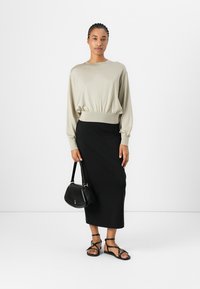 Beige gestrickter Pullover mit tailliertem Schnitt, kombiniert mit einem schwarzen, figurbetonten Midirock. Das Model trägt eine kleine schwarze Handtasche und schwarze Sandalen.