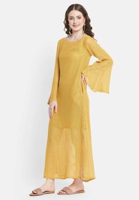 Gelbes, langärmeliges Maxikleid mit weitem Ärmeldesign und seitlichem Schlitz. Aus leichtem, durchscheinendem Material gefertigt. Kombiniert mit flachen Sandalen.