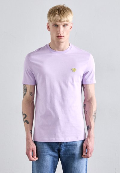 Lavender bomulds t-shirt med korte ærmer, rund halsudskæring og lille guld logo detalje på brystet. Båret med blå jeans.