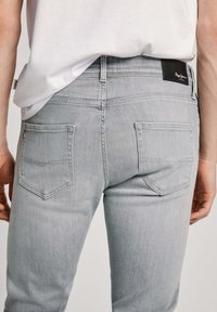 Sivi traperici slim kroja, s dva stražnja džepa i crnom logotip oblogom na pojasu. Glatka tekstura i krojeni dizajn.