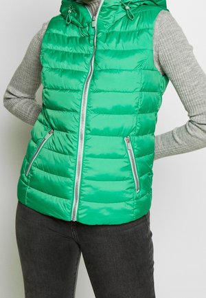 Personne portant une veste matelassée à capuche vert vif sur un pull gris côtelé à manches longues et un pantalon gris foncé, les mains posées sur les hanches.