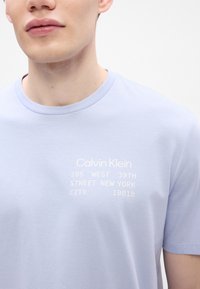 Hele sinine puuvilla T-särk lühikeste varrukatega. Kätkeb valge trükitud teksti: "Calvin Klein 205 WEST 39TH STREET NEW YORK CITY 10018". Ümmargune kaelus.