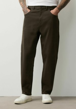 Man wearing dark brown straight-leg pants with side pockets, white sneakers, and a white shirt, standing against a plain background.