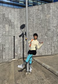 Sieviete atbalstā uz staba ar vienu kāju paceltu, valkājot gaiši zilas sportiskās bikses, svītrainas zeķes, baltas kedas un krēmkrāsas džemperi pret pelēka akmens sienas fona.