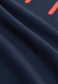 Tumesinine puuvillane kangas, millel on sile tekstuur, õrnad voldid ja silmatorkav koralloranž graafiline disainelement.