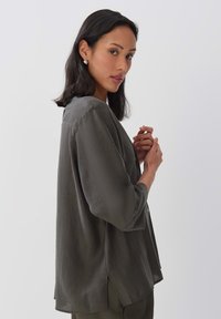 Blouse ample de couleur olive, en tissu texturé, avec des manches trois-quarts et des fentes sur les côtés pour faciliter les mouvements.