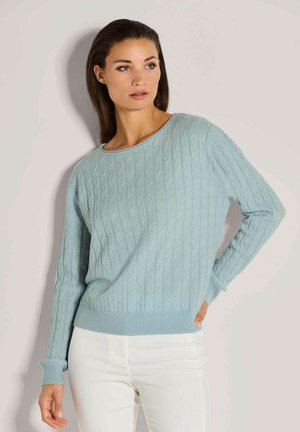 Frau mit langen dunklen Haaren, die einen hellblauen, strukturierten Pullover und weiße Hosen trägt, steht mit einer Hand an der Hüfte vor einer schlichten Wand.