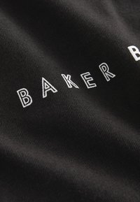 Črna tkanina z teksturirano površino, ki ima bele vezenine z napisom "BAKER". Oblikovanje je preprosto in čisto.