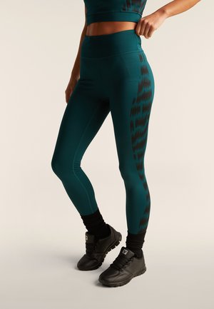 Leggings turquesas con una cintura alta, que presentan un patrón de zigzag negro en el costado. Textura mate con un acabado suave. Zapatillas de deporte negras.