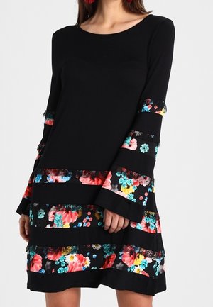 Donna che indossa un vestito nero a maniche lunghe con strisce orizzontali a stampa floreale in rosso, blu, giallo e rosa, con maniche svasate e scollo rotondo.