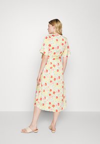 Vestido midi floral com mangas curtas e babados, uma cintura com laço, apresentando um fundo amarelo com flores vermelhas e rosas. Usado com sandálias slides bejes.