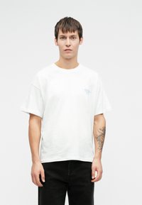 Mladenič s kratkimi rjavimi lasmi nosi navadno belo majico in črne hlače ter prikazuje tetovažo na svojem levem podlahtniku.