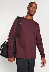 Burgunderfarget langermet skjorte laget av bomullsblanding, med en liten svart logo på brystet. Modellen bærer en svart treningsbag.