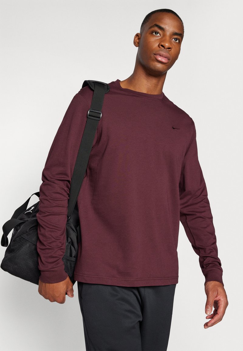 Burgundowa koszula z długim rękawem, wykonana z mieszanki bawełny, z małym czarnym logo na piersi. Model niesie czarną torbę sportową.