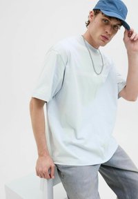 Helles blaues, übergroßes T-Shirt aus Baumwolle, mit kurzen Ärmeln und einem grafischen Druck. Kombiniert mit hellgrauen Jeans und einer blauen Kappe.