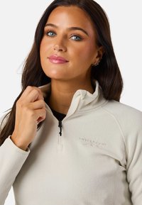 Lys beige fleece pullover med kvart lynlås og lynlåset krave samt broderet logo på brystet. Glat tekstur og tætsiddende ærmer.