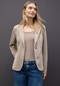 Beige korduroy sako s dvěma knoflíky, klopami a předními kapsami, nošené přes šedý top a modré džíny. Hladká textura.