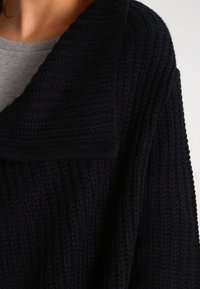 Cardigan noir en tricot avec un large col et un motif texturé. Le tissu semble doux et épais, idéal pour les superpositions.