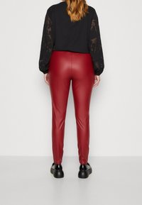 Røde faux læderbukser med slim fit. Glat tekstur med høj glans. Parret med en sort top med transparente blomsterærmer. Sorte sko.