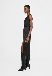 Lang zwart jurk met een zijsplit, gemaakt van stijlvolle stof, gecombineerd met zwart hoge laarzen met hakken tot de knie. Eenvoudig ontwerp en elegante silhouet.