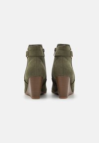 Bottines en daim vert olive avec un talon empilé marron, dotées d'une fermeture éclair latérale et d'une sangle décorative près de la cheville pour un détail supplémentaire.