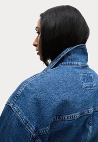 Giacca in denim blu con colletto alto e cuciture in vista. Presenta una patch rettangolare sulla parte superiore della schiena. Il tessuto ha una finitura testurizzata.