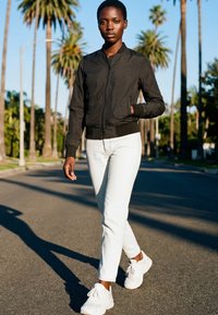 Jonge vrouw die op een met palmbomen omzoomde straat wandelt, gekleed in een zwarte bomberjack, witte broek en witte sneakers in fel daglicht.
