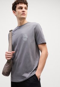Grijze katoenen T-shirt met een ronde hals, korte mouwen en een subtiel gedrukt logo op de borst. Geko gecombineerd met een grijze schoudertas.