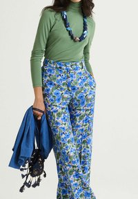 Maglia a maniche lunghe verde abbinata a pantaloni con stampa floreale multicolore, con tonalità di blu e verde. Accessori includono uno scialle blu e una borsa con nappe.