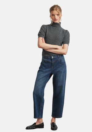 Frau mit blondem Haar, die einen grauen kurzärmligen Rollkragenpullover, blaue Jeans mit geradem Bein und schwarze Loafer trägt, steht mit verschränkten Armen.
