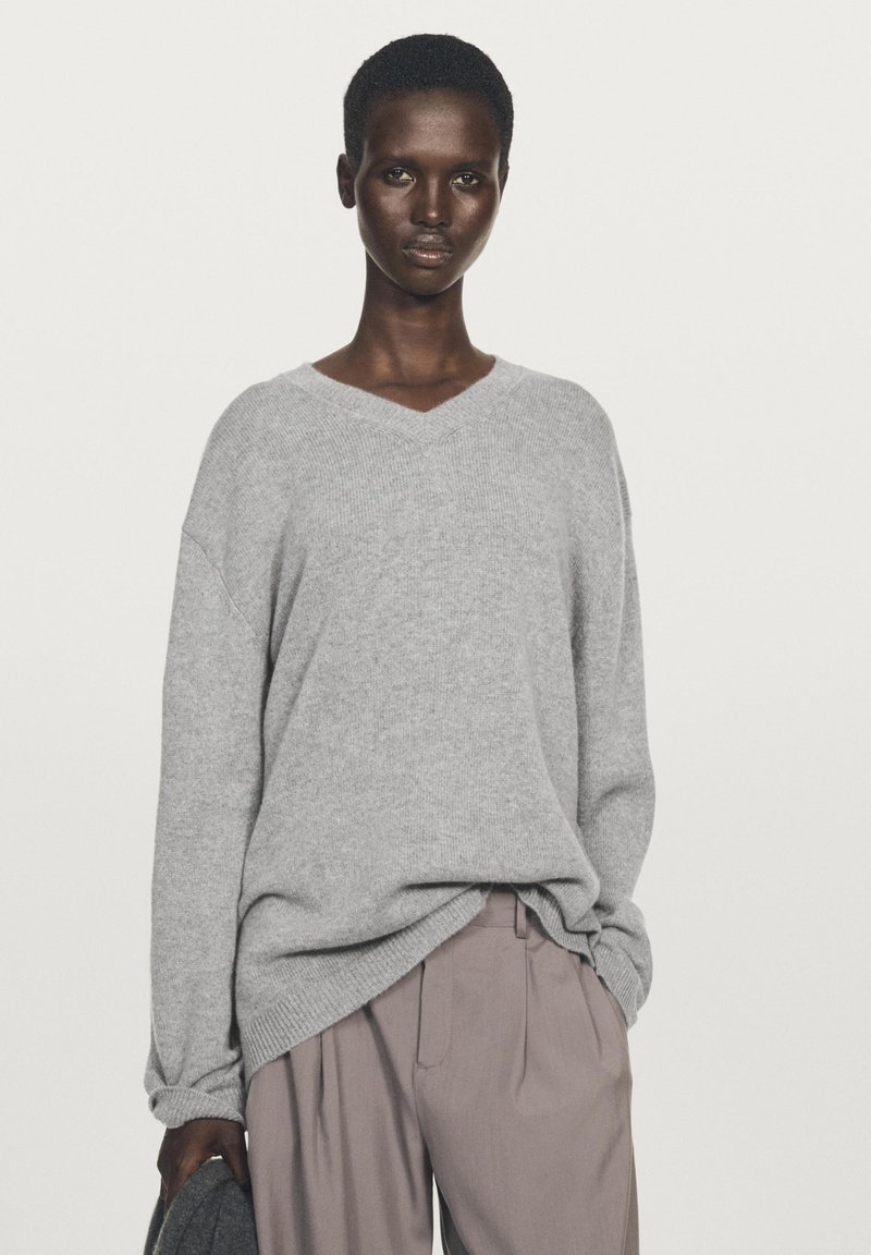 Grauer übergroßer V-Ausschnitt-Pullover aus weichem Strickmaterial, mit langen Ärmeln und lockerem Schnitt, kombiniert mit hellgrauen, plissierten Hosen.