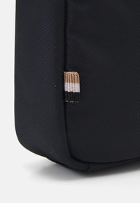 Angolo di una borsa di stoffa nera con una texture rigata. Presenta un piccolo etichetta tessuta con strisce nere, bianche e marroni. Forma rettangolare.