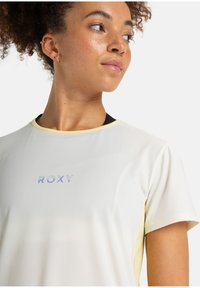Camiseta deportiva de color amarillo claro con mangas cortas, un cuello redondo y un sutil degradado. Presenta el logotipo "ROXY" en azul.