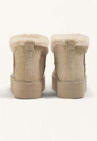 Beige mockaskängor med en fluffig krämfärgad pälskrage och en tjock plattformssula. Texturerat material med sömnadsdetaljer på sidorna.