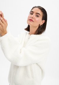 Veste blanche en peluche zippée avec un col haut et des manches surdimensionnées, présentant une texture douce et un design minimaliste. Pas de motifs visibles.