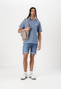 Grå crossover-veske, blå bomulls-T-skjorte, denimshorts, hvite sneakers med sorte striper. Modell står foran en helt hvit bakgrunn.