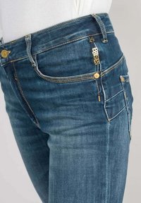 Jeans de denim en azul oscuro, con detalles dorados en los bolsillos y la cremallera, con un diseño slim fit y un ligero desgastado en la tela.