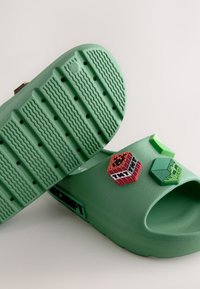 Sandales à glissière vertes en caoutchouc, avec semelles texturées et trois ornements 3D colorés : bloc TNT rouge, hexagone vert et un autre bloc texturé.